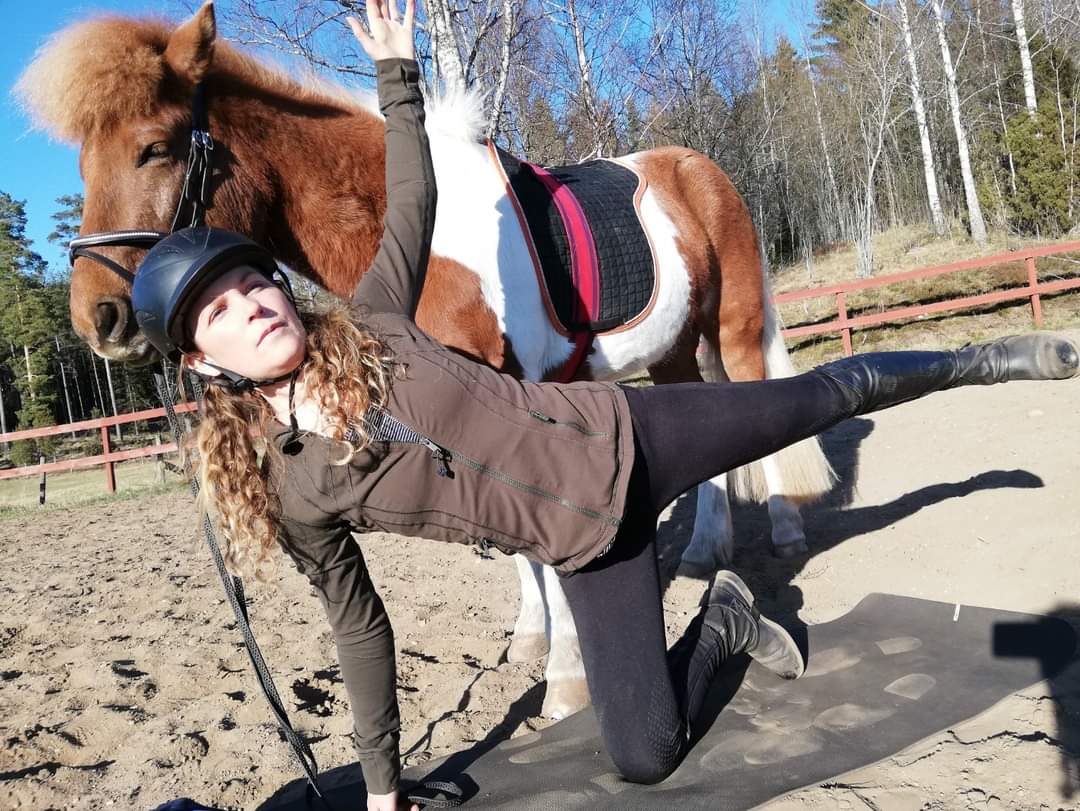 En tjej gör yoga på en ridbana med en häst i bakgrunden.